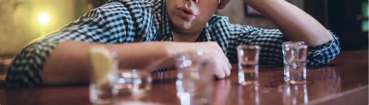 Person sitting at a bar with several empty shot glasses, appearing distressed.
