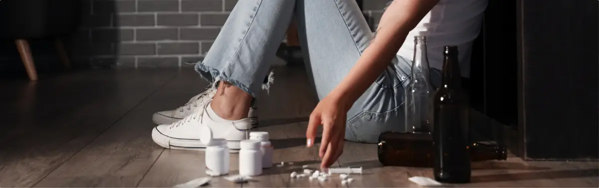 Person sitting on the floor near opiate prescription bottles, pills, and other ilicit substances.