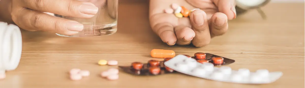 Hand holding assorted prescription pills beside a glass of water and blister packs on a table.